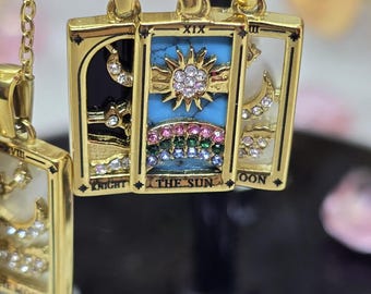 May include: Three gold-toned rectangular pendants on chains. The center pendant features a blue background with a gold sun, clear crystals, and a multi-colored crystal rainbow, labeled THE SUN XIX. Flanking pendants show moons and crystals.