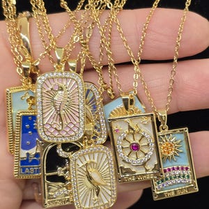 May include: A set of gold necklaces with colorful enamel pendants featuring tarot card imagery. The pendants are rectangular and have a gold frame with clear rhinestones. The pendants feature images of the Sun, the Wheel, the Knight of Swords, the Last Judgement, and the High Priestess.