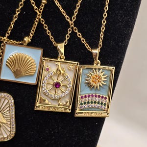 May include: Three gold necklaces with pendants featuring tarot card designs. The pendants are rectangular and have a white background with colorful details. The pendants feature the cards "The Wheel", "The Sun", and a third card with a seashell design.