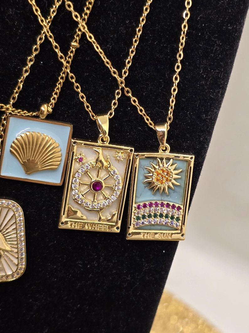 May include: Three gold necklaces with pendants featuring tarot card designs. The pendants are rectangular and have a white background with colorful details. The pendants feature the cards "The Wheel", "The Sun", and a third card with a seashell design.