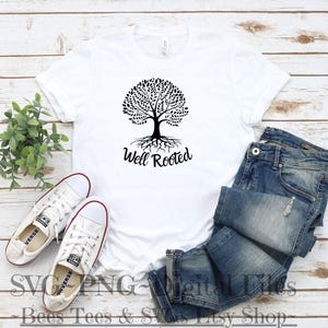 May include: White t-shirt with a black tree design and the text "Well Rooted". The t-shirt is laying on a wooden surface with a pair of blue jeans and a pair of white sneakers.