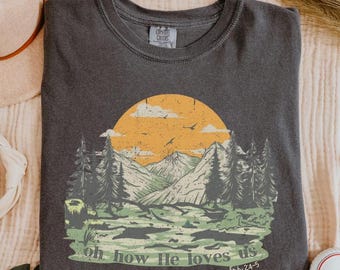 Op de afbeelding: Een donkergrijs T-shirt met een afbeelding van een bergketen met een gele zon en de tekst "oh how He loves us Eph 2:4-5".