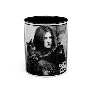 May include: White ceramic mug with a black interior and a black and white graphic. The graphic shows a person with long hair holding a black cat, with the words "HOWL N HISS" in a stylized font.