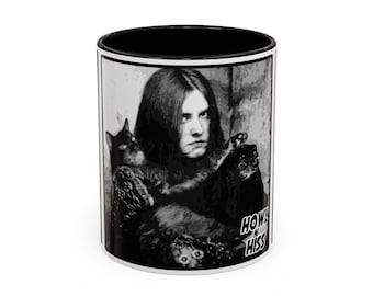 May include: White ceramic mug with a black interior and a black and white graphic. The graphic features a person with long hair holding a black cat, with the words "HOWL N HISS" in a stylized font.