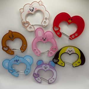 May include: A collection of seven colorful, heart-shaped carabiner clips. Each clip features a different cartoon character design, including a sheep, bunny, koala, and bear. The clips are made of translucent plastic and have a metal clasp.