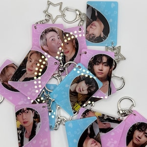 May include: A collection of rectangular acrylic keychains featuring portraits of various individuals. The keychains are colored in shades of light pink and light blue, decorated with small white heart and musical note patterns, and attached to silver star-shaped metal clasps.