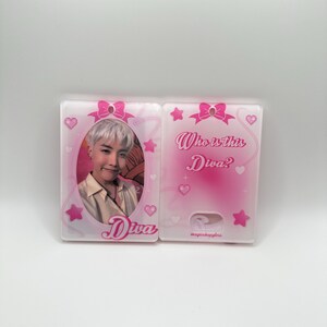 May include: A pink and white plastic charm with a photo of a person with light hair and a white shirt. The charm has a pink bow and stars. The text "Who is this Diva?" and "Diva" are printed on the charm.