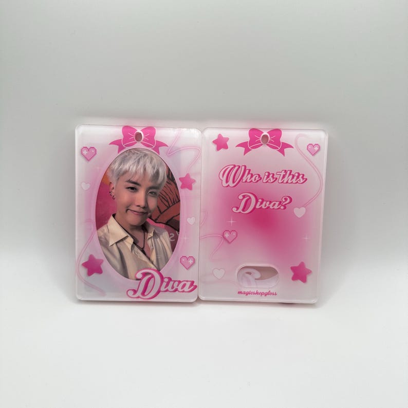 May include: A pink and white plastic charm with a photo of a person with light hair and a white shirt. The charm has a pink bow and stars. The text "Who is this Diva?" and "Diva" are printed on the charm.