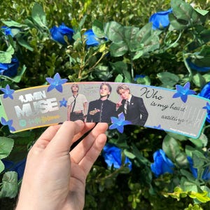 May include: A silver and holographic coffee sleeve with the text "#UMN MUSE" and a Spotify code. The sleeve features a photo of three men with the text "Love u... Jimin" and "Who is my heart waiting for?" The sleeve is decorated with blue flowers.