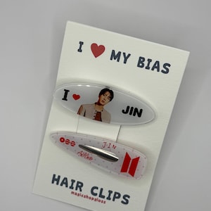 May include: Two oval hair clips on a white display card labeled I LOVE MY BIAS and HAIR CLIPS. The top clip features a photo of Jin with a red heart. The bottom clip is translucent with white dots, red text reading JIN and ARIRANG, and a red logo.