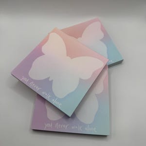 May include: Square notepads with a pastel gradient background of pink, purple, and blue. Each notepad features a white butterfly silhouette and the handwritten text "you never walk alone".