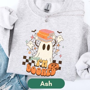 Può includere: Felpa grigia con grafica a tema Halloween. Il design presenta un fantasma che tiene dei libri, circondato da pipistrelli, stelle e una ragnatela. Il testo "Sorry I'm Booked" è visualizzato con una zucca e un motivo a scacchi.
