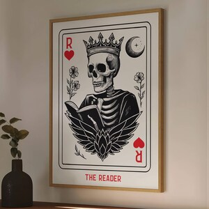 Può includere: Stampa artistica incorniciata raffigurante uno scheletro con una corona che legge un libro, progettata in stile carta da gioco. La stampa include il testo "THE READER" e simboli di cuori rossi.