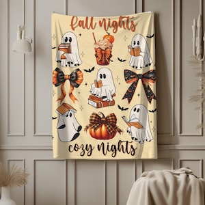 Può includere: Una coperta color crema a tema Halloween. Presenta fantasmi che leggono libri, zucche, fiocchi e pipistrelli. Le parole "Fall nights" e "cozy nights" sono scritte sulla coperta.