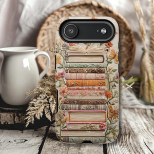 Può includere: Una custodia per telefono con un design di libri impilati, adornata con fiori e farfalle. La custodia ha uno sfondo color crema con fiori e farfalle colorati. I libri sono in vari colori, tra cui arancione, rosa e verde.
