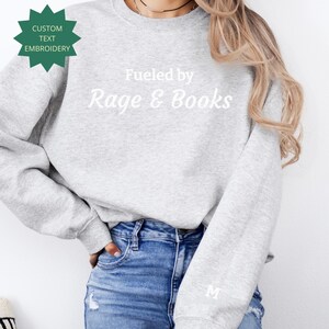 Può includere: Una felpa grigio chiaro con la scritta bianca "Fueled by Rage & Books". La felpa ha anche la scritta "Custom Text Embroidery" a forma di stella verde. Una lettera "M" bianca è sulla manica.