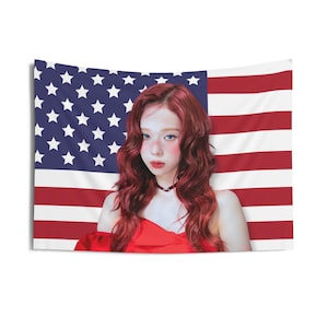 Peut inclure: Une femme aux longs cheveux roux ondulés se tient devant un drapeau américain. Elle porte une robe rouge sans bretelles et un collier ras du cou noir.