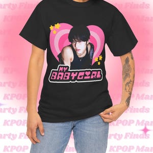 May include: Black t-shirt with a pink heart graphic featuring a person with black hair and a silver necklace. The text "MY BABYGIRL" is printed in pink and white letters on the shirt.