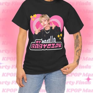 Può includere: T-shirt nero con grafica di una persona con capelli biondi, un cuore rosa e la scritta "MY BABY GIRL". Il design include stelle gialle. La maglietta è indossata con jeans blu.