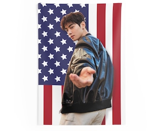 May include: A man wearing a black leather jacket and white trousers stands in front of an American flag. He is looking at the camera and reaching out with his right hand.