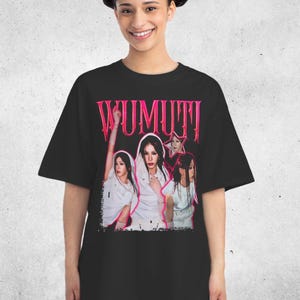 May include: Black t-shirt featuring a graphic of three women in white outfits with the word "WUMUTI" in pink, gothic-style lettering. The image has a vintage, distressed aesthetic.