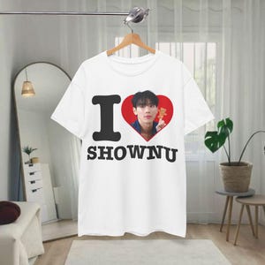 May include: A white short-sleeved t-shirt featuring a graphic print with the black text I, a red heart containing a photograph of a person holding a small brown bear, and the black text SHOWNU positioned directly below the heart graphic.