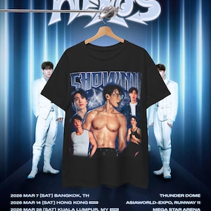 May include: A black short-sleeve t-shirt featuring a graphic print of the artist Shownu in multiple poses, including a shirtless central image. The text "SHOWNU" appears in a metallic-style font above the images, set against a dark blue background.
