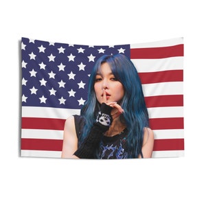 May include: A woman with blue hair is posing in front of an American flag. She is wearing a black shirt and has her finger to her lips.