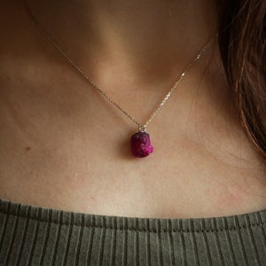 Può includere: Una collana d'argento con un pendente di pietra preziosa grezza color fucsia. Il pendente ha una forma irregolare ed è sospeso a una catena sottile. La collana è mostrata su uno sfondo neutro.