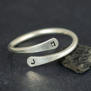 May include: A silver-colored wrap ring with the initials "J" and "M" stamped on the ends. The ring is displayed on a dark surface next to a small, dark rock.