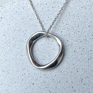 Pode incluir: Um colar prateado com um pendente de círculo irregular e liso. O pendente pende de uma corrente fina. A joia é exibida sobre uma superfície clara e texturizada, criando uma estética minimalista.