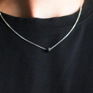 May include: A silver chain necklace featuring a small, dark bead pendant. The necklace is set against a black top, providing contrast. The jewellery is simple and minimalist in design.