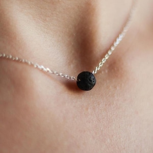 Può includere: Una delicata collana d'argento con un piccolo pendente rotondo in pietra lavica nera. Il pendente è centrato su una sottile catena d'argento, creando un design minimalista ed elegante. La collana è indossata intorno al collo.