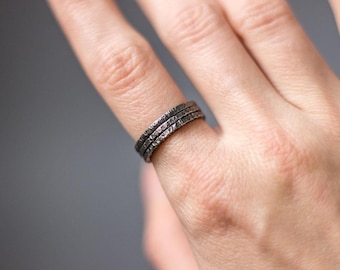 May include: Three textured, dark grey rings are stacked on a finger. The rings have a rough, uneven surface, giving them a handcrafted look. The jewellery is likely made of metal and has a rustic appearance.