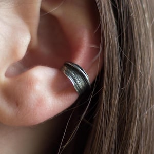 Op de afbeelding: Een zilveren oormanchet met een getextureerd, gelaagd ontwerp. De manchet omsluit het kraakbeen van het oor, waardoor een moderne, minimalistische look ontstaat. Het metaal heeft een licht geoxideerde afwerking.