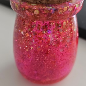Op de afbeelding: Een kleine, ronde, roze glazen pot met een kurken deksel. De pot is gevuld met roze glitter en iriserende glitter.