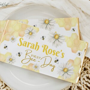 Può includere: Un involucro di tavoletta di cioccolato stampabile bianco e giallo con un motivo a nido d'ape e margherite. Il testo sull'involucro recita "Sarah Rose's First Bee-Day".