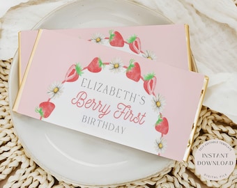 May include: Two pink candy bar wrappers with gold edges, featuring a strawberry and daisy design. The text reads "Elizabeth's Berry First Birthday". The wrappers are on a white plate.