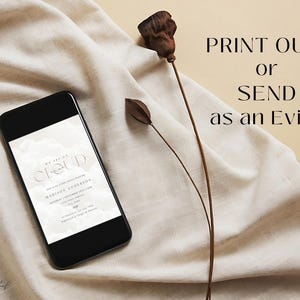 May include: A digital invitation on a smartphone screen with a white background and the text "We are on a Cloud" and "Madison Anderson". The text is in a modern, minimalist font. The phone is lying on a beige fabric surface with dried flowers.