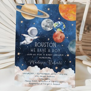 May include: A blue and white baby shower invitation with a watercolor illustration of an astronaut holding planets. The text reads "Houston, We Have A Boy" and includes the baby's name, Madison Roberts, the date and time of the shower, and RSVP information.