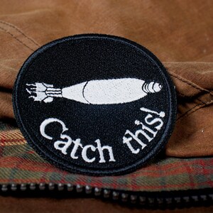 May include: Black and white embroidered patch with a cartoon image of a rocket and the text "Catch this!"