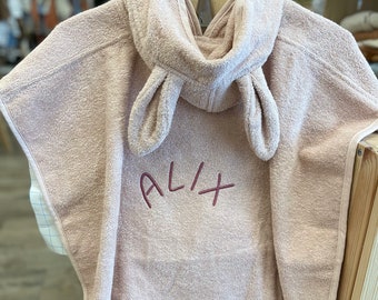 May include: A light pink hooded towel with bunny ears and the name "Alix" embroidered on the front. The towel is hanging on a wooden hanger.
