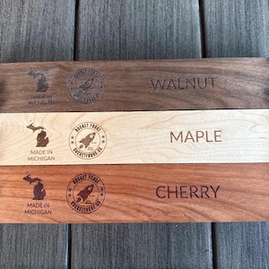 May include: Three wooden planks labeled Walnut, Maple, and Cherry, each with a different wood grain and color. Each plank has the text "Made in Michigan" and a rocket logo. The planks are held together with black hardware.