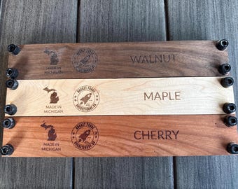 May include: Three wooden planks labeled Walnut, Maple, and Cherry, each with a different wood grain and color. Each plank has the text "Made in Michigan" and a rocket logo. The planks are held together with black hardware.