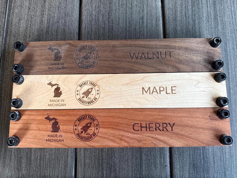 May include: Three wooden planks labeled Walnut, Maple, and Cherry, each with a different wood grain and color. Each plank has the text "Made in Michigan" and a rocket logo. The planks are held together with black hardware.