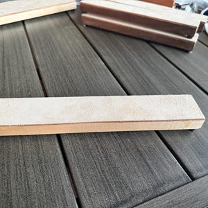 May include: A rectangular sharpening stone with a light tan suede-like surface and a wooden base. Several other similar stones are visible, suggesting a set or related items. The items are on a gray wooden surface.