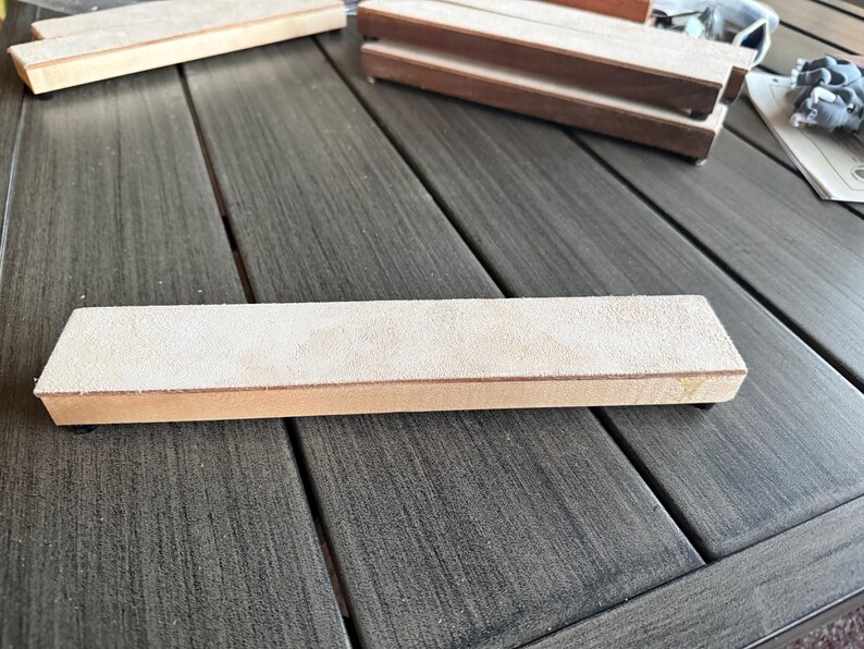 May include: A rectangular sharpening stone with a light tan suede-like surface and a wooden base. Several other similar stones are visible, suggesting a set or related items. The items are on a gray wooden surface.