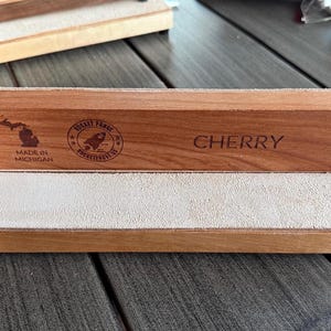 May include: A cherry wood product with a light-colored leather surface. The wood is engraved with the word "CHERRY", a rocket logo, and "MADE IN MICHIGAN". Black hardware secures the wood to the base. The product is displayed on a wooden surface.