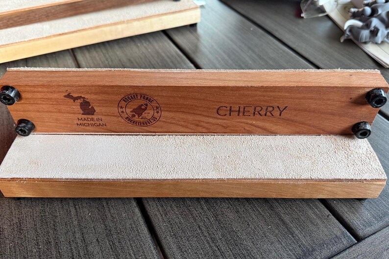 May include: A cherry wood product with a light-colored leather surface. The wood is engraved with the word "CHERRY", a rocket logo, and "MADE IN MICHIGAN". Black hardware secures the wood to the base. The product is displayed on a wooden surface.