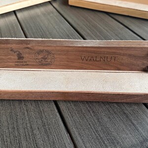 May include: A walnut wood desk organizer with a light-colored suede interior. The organizer has the word "WALNUT" engraved on the front. The text "Made in Michigan" and a logo are also visible. Black screws secure the wood.